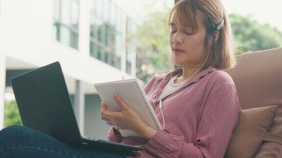 年轻的亚洲女孩戴着耳机用笔记本电脑上网学习躺在游泳池附近的椅子上写笔记本