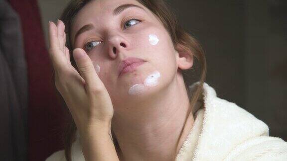 迷人的年轻白人女子在浴室里对着镜子往脸上抹面霜面部皮肤护理按摩的动作