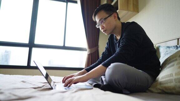 年轻人坐在床上在家里的卧室里用笔记本电脑工作