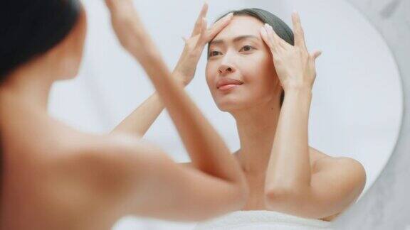 美丽的亚洲妇女肖像轻轻应用面霜在浴室镜子年轻的成年女性使她的皮肤柔软光滑与天然保湿化妆品护肤品