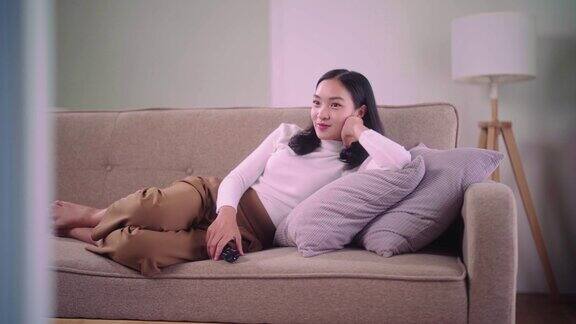 年轻的亚洲女人拿着遥控器坐在家里客厅舒适的大沙发上一边看电视一边吃爆米花