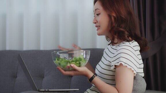 亚洲年轻女子看着笔记本电脑快乐地在家里吃着新鲜的沙拉