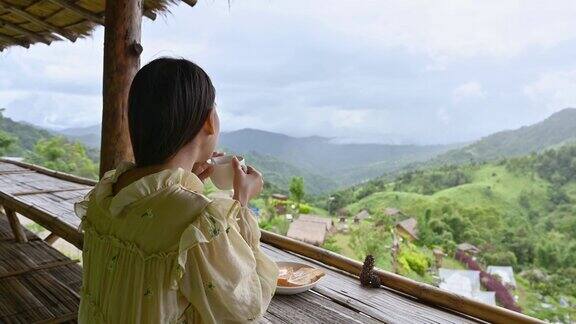 年轻的亚洲女人喝热巧克力和吐司在小屋阳台之间的山早上