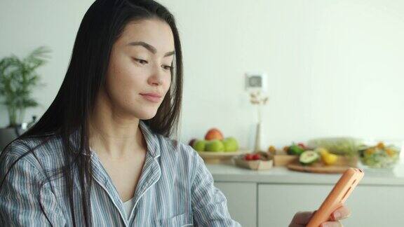 亚洲女孩穿着睡衣喝果汁在家里的厨房使用智能手机触摸屏