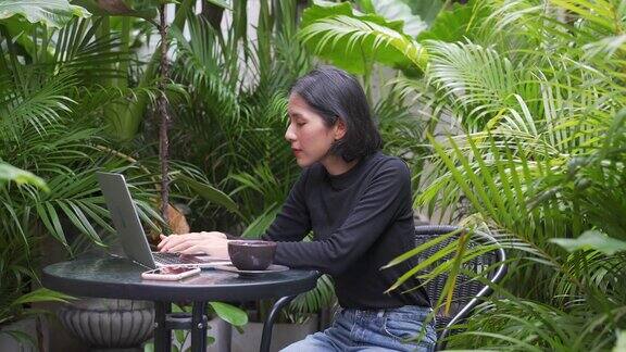 4K亚洲妇女在笔记本电脑上工作喝着咖啡在花园里