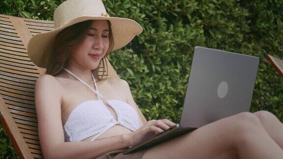 亚洲女性在度假期间躺在游泳池旁的椅子上使用笔记本电脑
