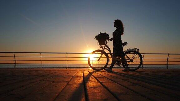 年轻女子的剪影穿着复古自行车和鲜花走在海边的木堤岸在日出或日落浪漫的旅行概念美丽的景色