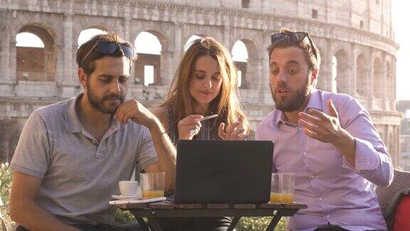 三个年轻人一起用笔记本电脑和平板电脑做一个项目头脑风暴写作交谈和研究坐在酒吧餐厅的桌子前在日落的罗马斗牛场