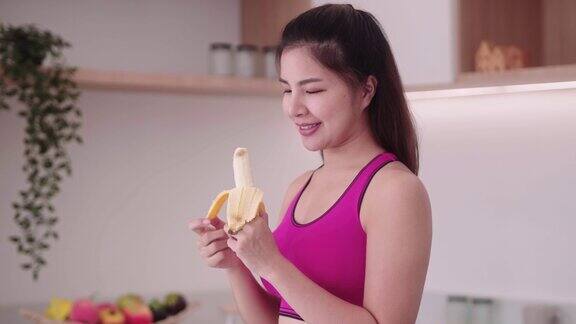 运动的年轻亚洲女性穿着运动服在厨房内吃香蕉享受吃水果的饮食和健康的运动减肥概念