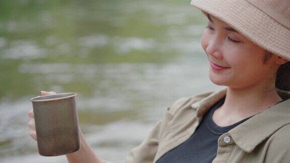 快乐的亚洲女人早上坐在河边喝咖啡在大自然中露营她感到放松和清爽休闲旅行和户外活动的生活方式