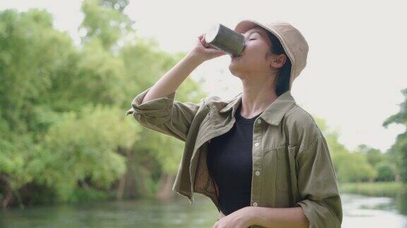 快乐的亚洲女人早上站在河边喝咖啡在大自然中露营她感到放松和清爽休闲旅行和户外活动的生活方式