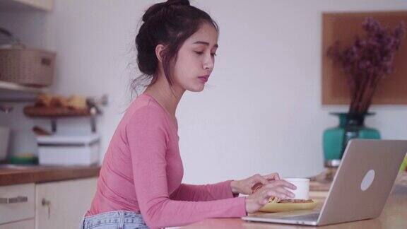 年轻快乐的亚洲自由职业女性早餐吃牛角面包喝咖啡在厨房柜台上用笔记本电脑工作享受在家工作