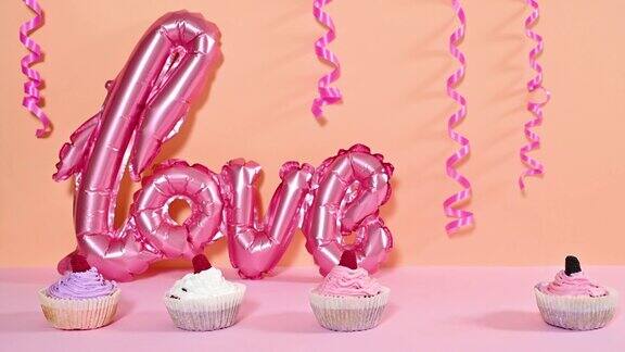 可爱的奶油杯形蛋糕出现在橙粉色粉彩主题上后面有丝带和爱心气球停止运动