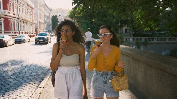 两个年轻的女孩穿着休闲装戴着太阳镜一边吃着冰淇淋一边微笑着聊天走在城市的街道上慢动作