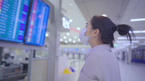 年轻的亚洲女性戴着白色防护口罩看机场航班时间安排显示屏在机场航站楼冠状病毒新冠肺炎大流行新常态距离病毒传播
