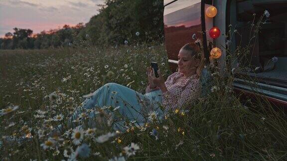 黄昏时分一名女子躺在停在草地上的一辆露营车旁一边使用智能手机