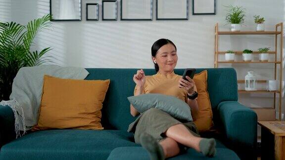 亚洲女性坐在家里客厅的沙发上戴着耳机和智能手机听音乐