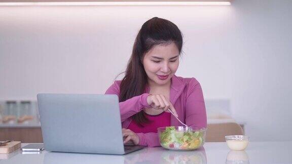 穿着运动服的年轻亚洲女性看着笔记本电脑在厨房里吃着蔬菜沙拉健康的饮食和运动的理念