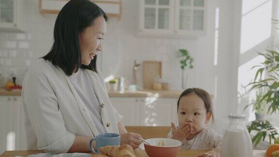 年轻的亚洲女人在家里的厨房里喂可爱的小孩笑