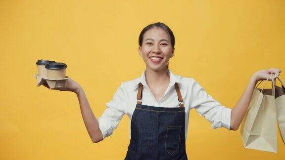 快乐的微笑亚洲女服务员穿着围裙友好地看着镜头给顾客咖啡或茶和美味的食品在手工纸袋隔离在黄色背景