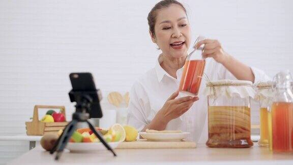 亚洲资深女性影响者教授如何制作自制的发酵生康普茶与不同的口味健康的天然益生菌口味饮料学习使用科技在厨房用智能手机与观众交谈