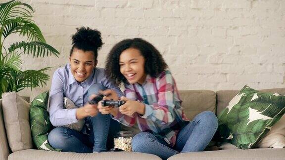 两个混合种族卷曲的女孩朋友坐在沙发上玩控制台电脑游戏与手柄有乐趣在家里