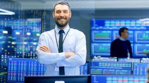 成功的男性股票交易员双臂交叉对着镜头微笑在繁忙的股票交易办公室交易员经纪人和交易商买卖债券显示器显示数字