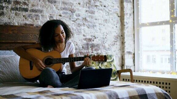 卷曲的非洲裔美国少女集中学习弹吉他使用笔记本电脑坐在家里的床上