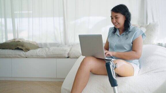 快乐的亚洲女性戴着假腿用笔记本电脑打字在网上聊天浏览网上冲浪社交媒体或在家在线工作腿部假肢设备截肢者概念