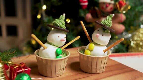 照片:在一间装饰着圣诞树的公寓里有两个自制的冰霜雪人杯形蛋糕用冰霜球做的黑巧克力饼干绿色奶油做的帽子粉色的鼻子和巧克力球做的绿色纽扣椒盐卷饼棒做的手臂