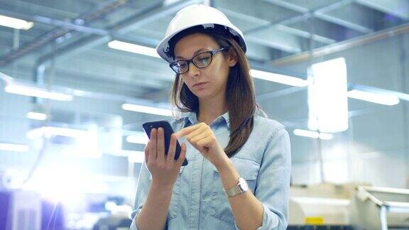 戴安全帽的女工业工程师站在大工厂里使用智能手机