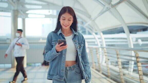 快乐的年轻亚洲女性用手机与朋友在线聊天女性穿着牛仔夹克走在城市使用智能手机上的社交媒体科技