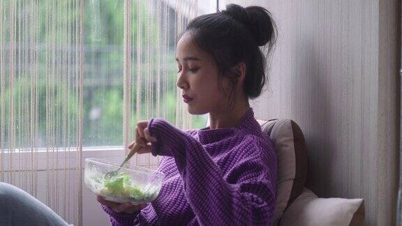 亚洲健康妇女拿着沙拉碗吃干净的蔬菜作为午餐在减肥饮食和健康的窗口附近的家庭饮食和健康食品