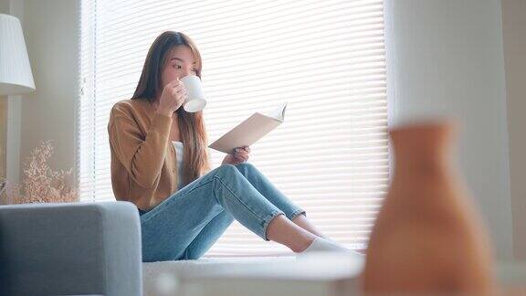 快乐的亚洲女人喝咖啡或茶在家里的客厅窗前看书放松生活方式的家