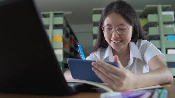亚洲大学生女孩读书工作和网上购物与笔记本电脑和智能手机在公共图书馆做她的家庭作业