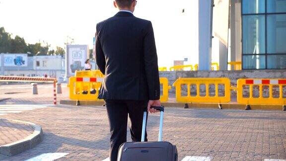 多莉拍摄了一个自信的年轻人穿着正式的黑色西装和他的行李走在城市的街道上在日落时分成功的商人去机场拖着行李箱近距离