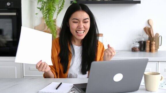 惊讶的亚洲女孩穿着时尚的休闲服装自由职业者IT专家坐在厨房的桌子上拿着笔记本电脑享受大利润庆祝胜利好消息用拳头做手势开心地微笑