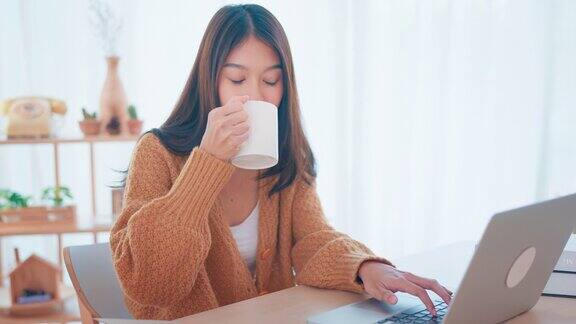 微笑的年轻亚洲女子在家里端着一杯咖啡使用笔记本电脑生活方式