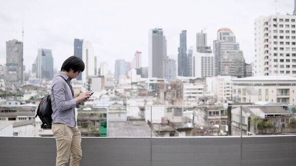 年轻的亚洲商人在办公大楼屋顶露台上使用智能手机背景是城市景色在智能设备上使用社交媒体应用程序或玩游戏