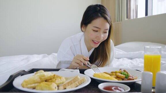 画像美丽的年轻亚洲女人穿着浴袍享受早餐在卧室的床上