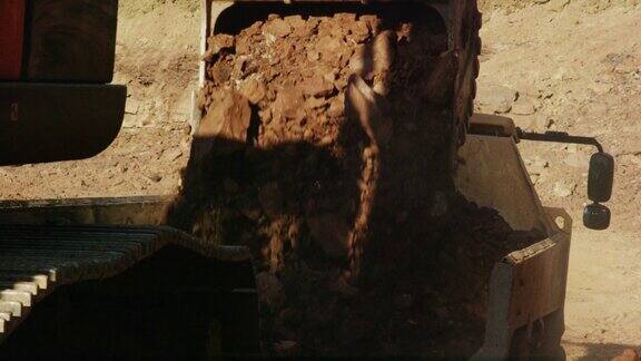 在阳光明媚的建筑工地挖掘机的铲斗向自卸车后面倾倒石块和泥土的慢动作镜头(铰接式自卸车)
