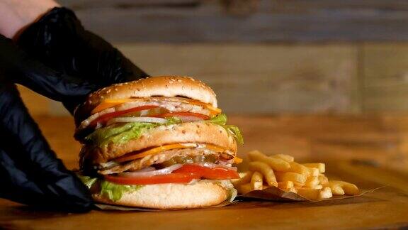 黑手套把一个多汁的汉堡、两片肉片和蔬菜放在薯条旁边的木板上不健康的快餐