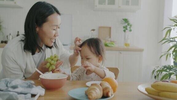 可爱的孩子吃羊角面包时慈爱的母亲带着葡萄在厨房在家里的慢动作肖像