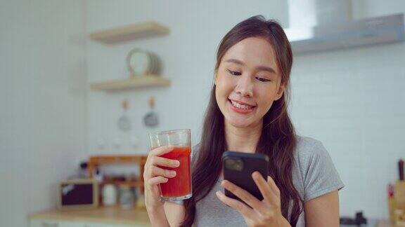 年轻的亚洲女人在家里的厨房里享受着果汁和电话