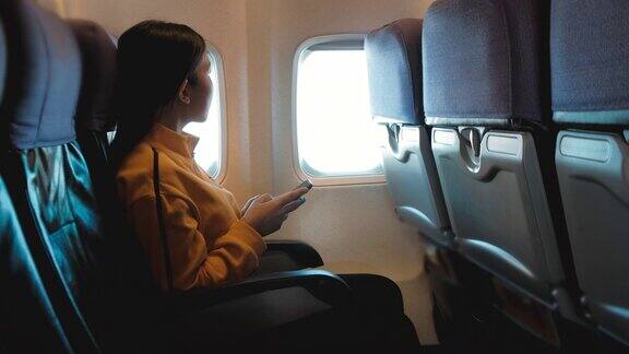 亚洲女游客坐在飞机窗口附近在飞行中使用智能手机