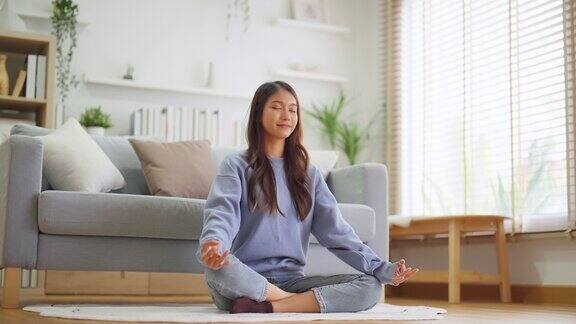 快乐的年轻亚洲女性在家里练习瑜伽和冥想坐在客厅的地板上以莲花的姿势闭上眼睛放松正念冥想和幸福概念