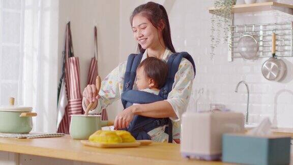 快乐的新亚洲妈妈抱着婴儿在前面的婴儿袋和做早餐微笑的妈妈抱着小婴儿婴儿准备健康的食物妈妈和宝宝在厨房共度时光
