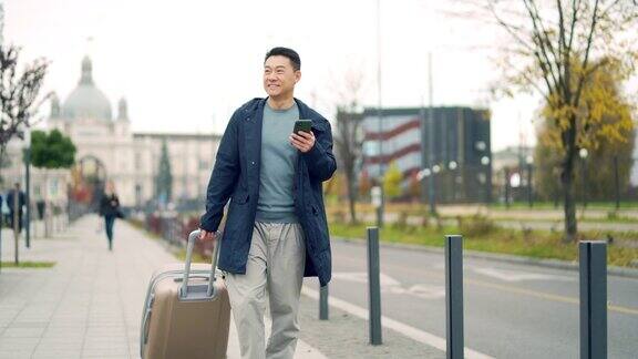 亚洲男性游客拿着包或拉着行李箱走在城市街道上使用手机一名男子来到一个新的欧洲城镇他走在人行道上使用智能手机