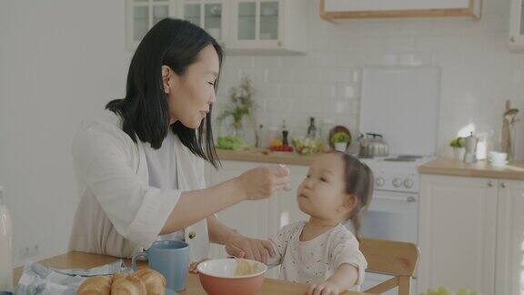 快乐的亚洲女士用婴儿食品喂小女儿微笑地看着公寓厨房里的孩子