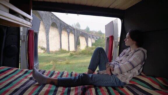 女人坐在房车的床上看着老桥的风景